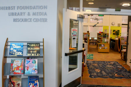 A Display Rack Holding Six Children’s Books Outside The Entrance To The Herbst Foundation Library &Amp;Amp; Media Resource Center, With A View Into The Library Interior Featuring Bookshelves And A Patterned Area Rug.