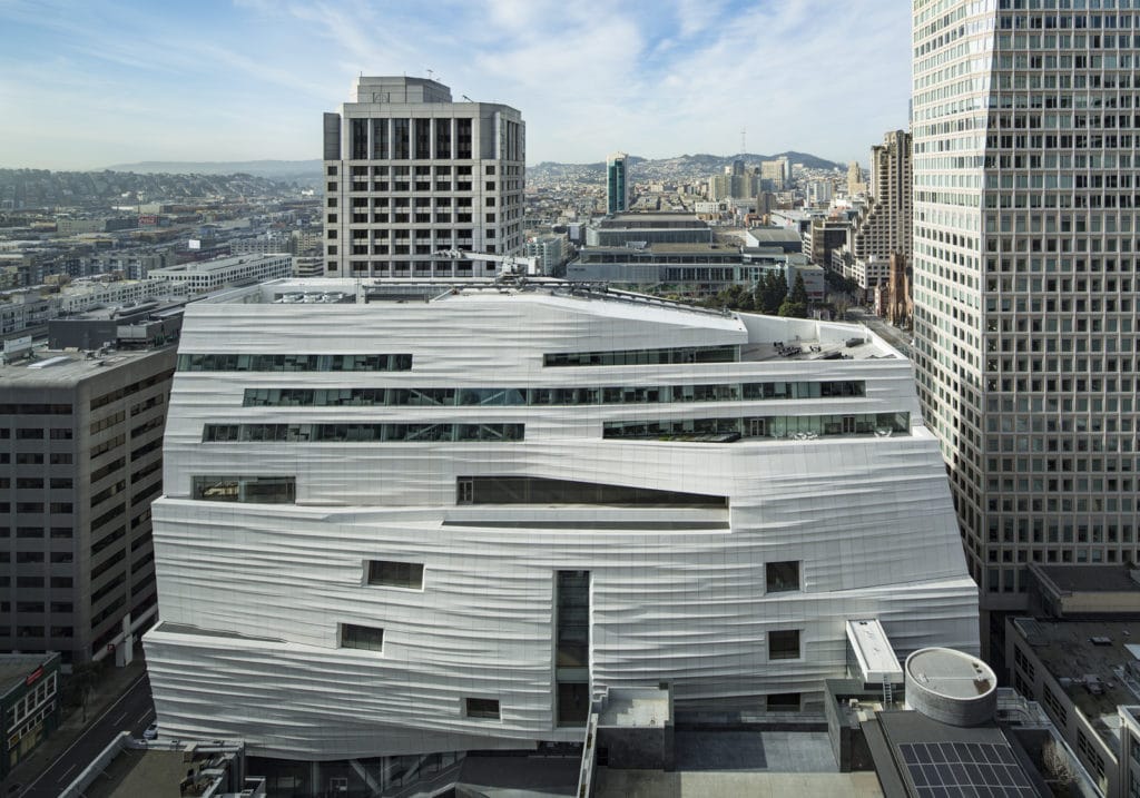 High-angle view of the white, wavy-textured facade of the SFMOMA building surrounded by city skyscrapers.