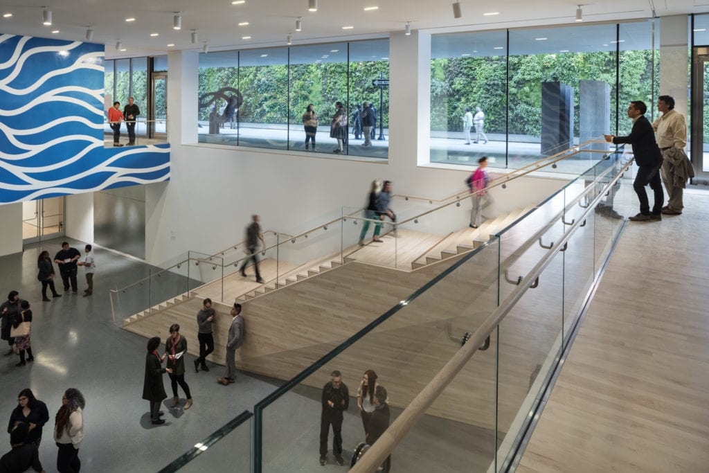 People gather in a bright, modern lobby with a wide wooden staircase, a wall mural featuring blue waves, and large windows looking out onto a green wall.
